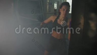 慢动作射击：强壮的女踢脚手在黑暗的健身房里用拳击袋运动，带着烟雾，踢着一